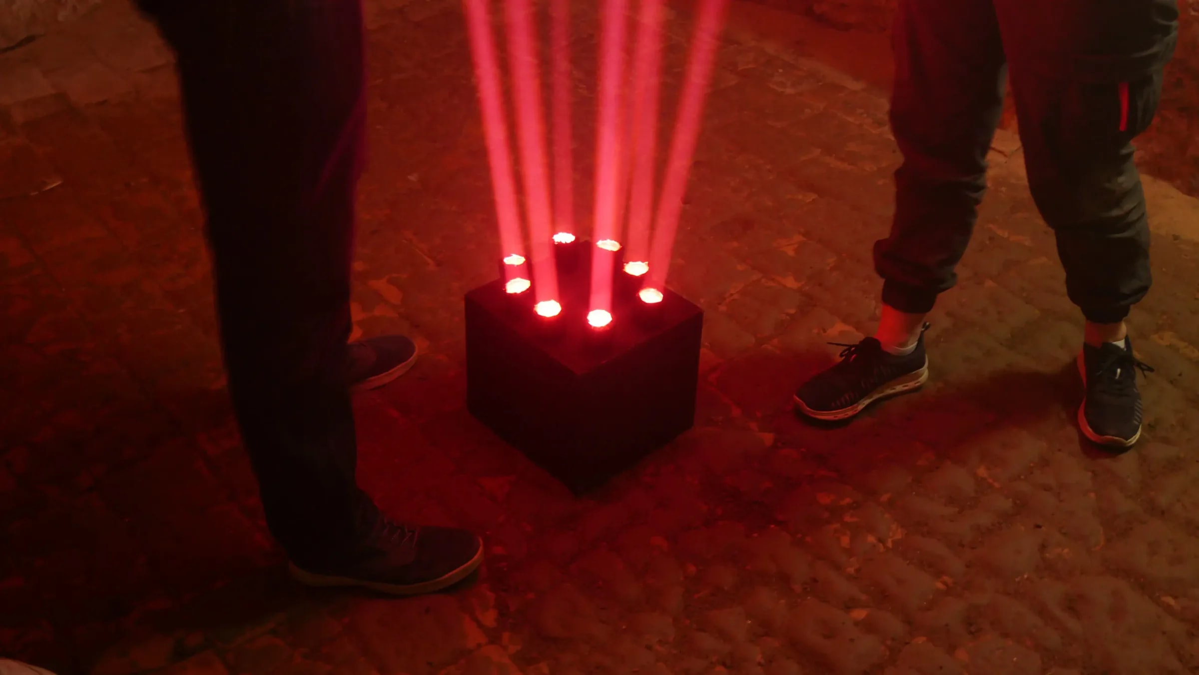 Sechs rote Lichtstrahlen steigen kreisförmig aus einem Podest auf, zwei Besuchende stehen daneben