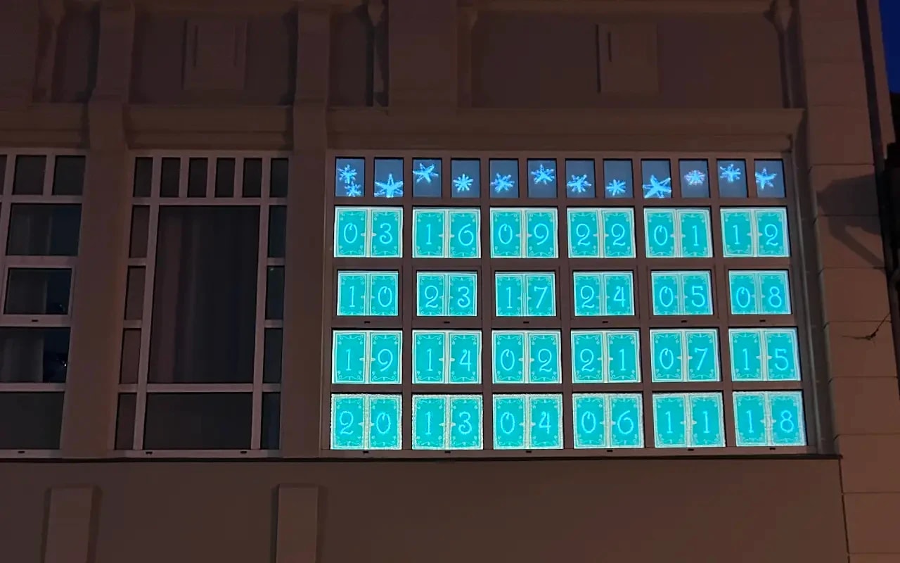 All 24 doors of the projected advent calendar glow turquoise in the windows of the department store, with a row of snowflakes above.