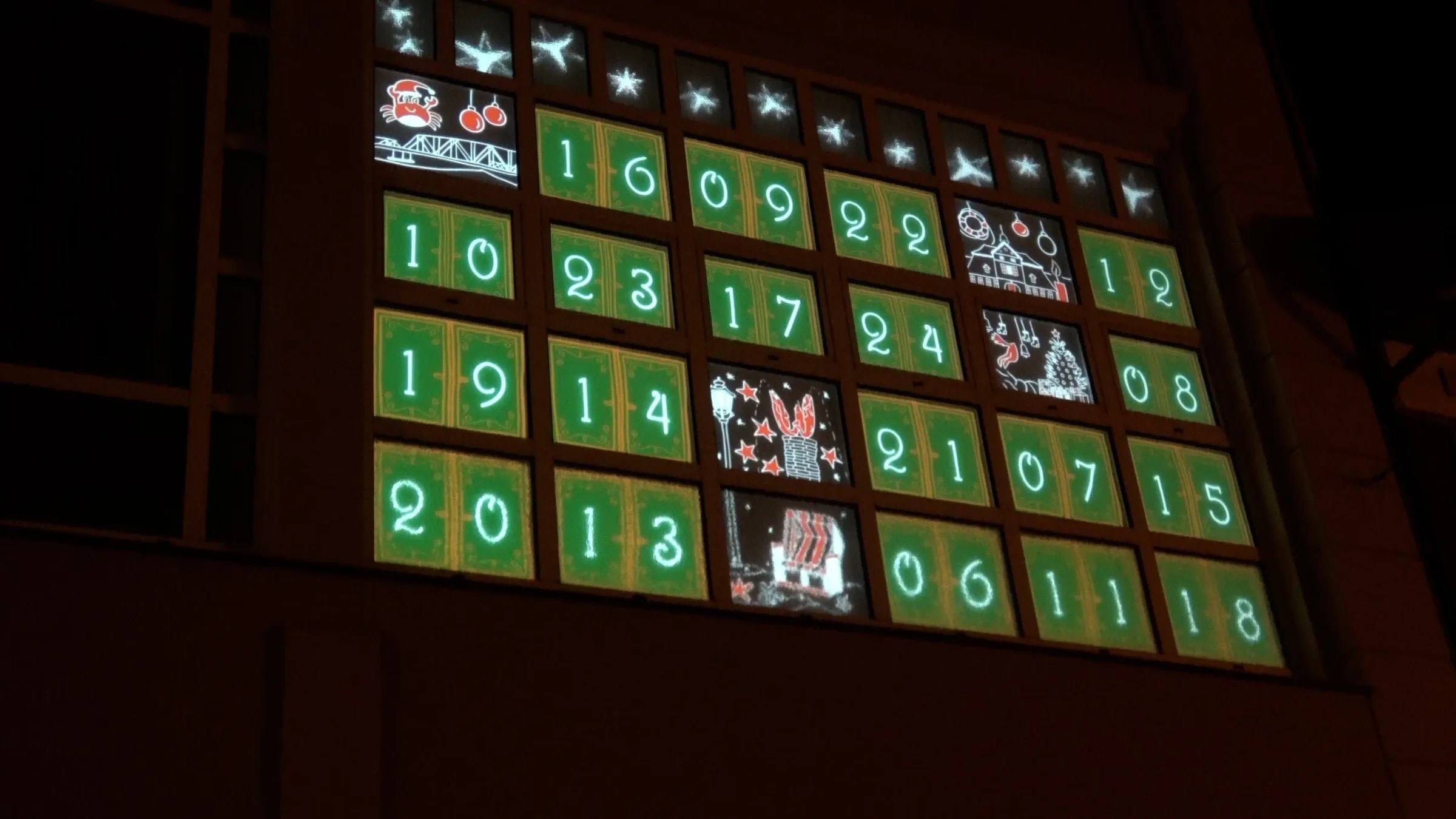 The facade of the old department store in Barth at night: some windows already show their motif while the rest still carry green numbered doors.