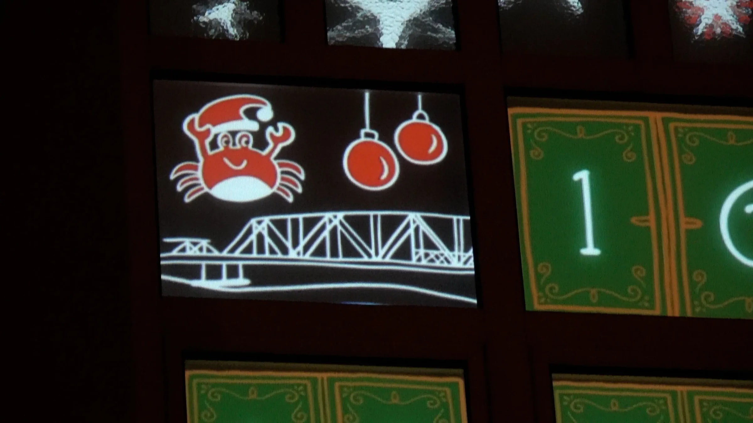 Close-up of the window projection in Barth: a crab wearing a Santa hat above a steel truss bridge, with two red baubles beside it and an unopened calendar door alongside.