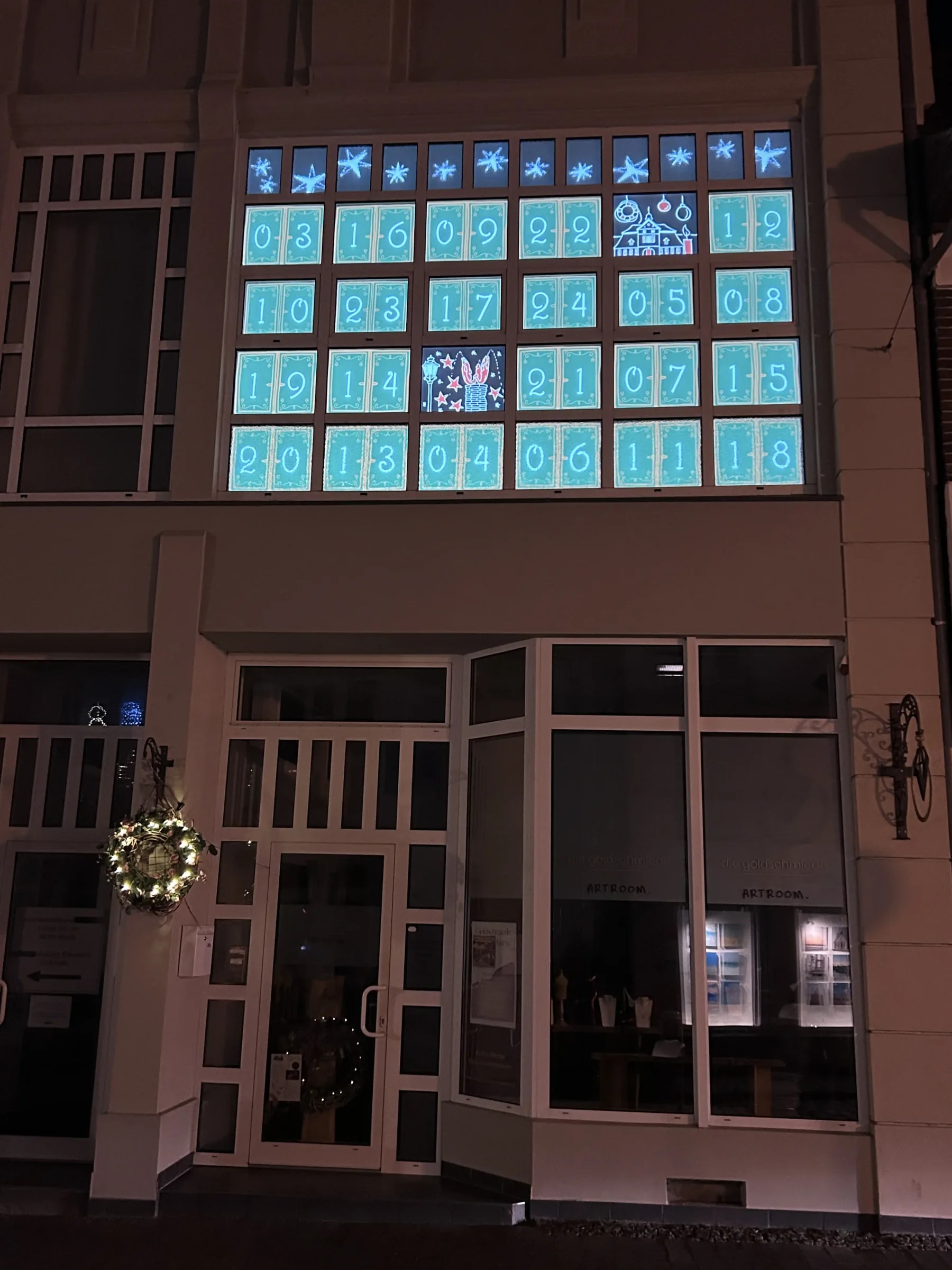 Façade in Barth at night with a projected advent calendar: turquoise numbered doors, two already opened, glowing stars above.