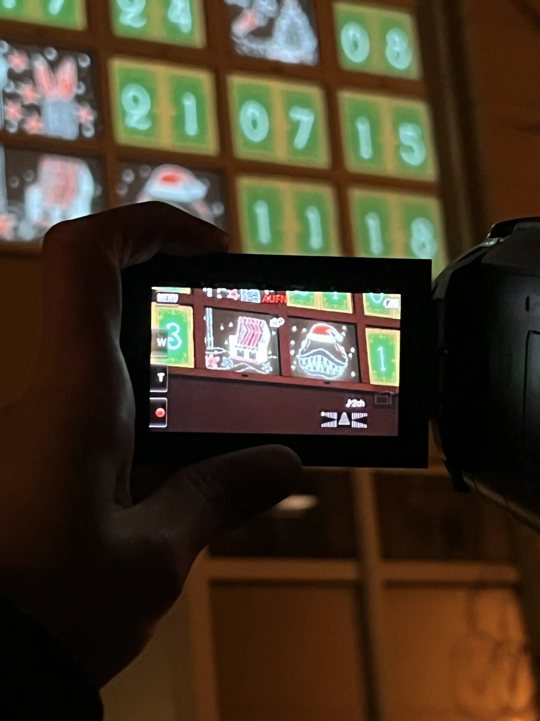 Video camera filming the advent calendar projection, its display showing a beach chair and Santa hat, green numbered doors glowing behind.