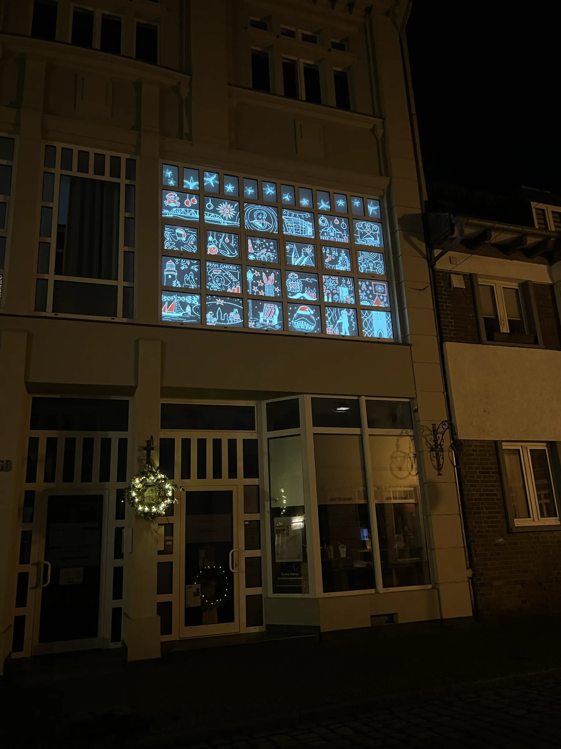 Period façade in Barth at night, all 24 windows shown as opened advent calendar doors with glowing motifs in blue, white and red.