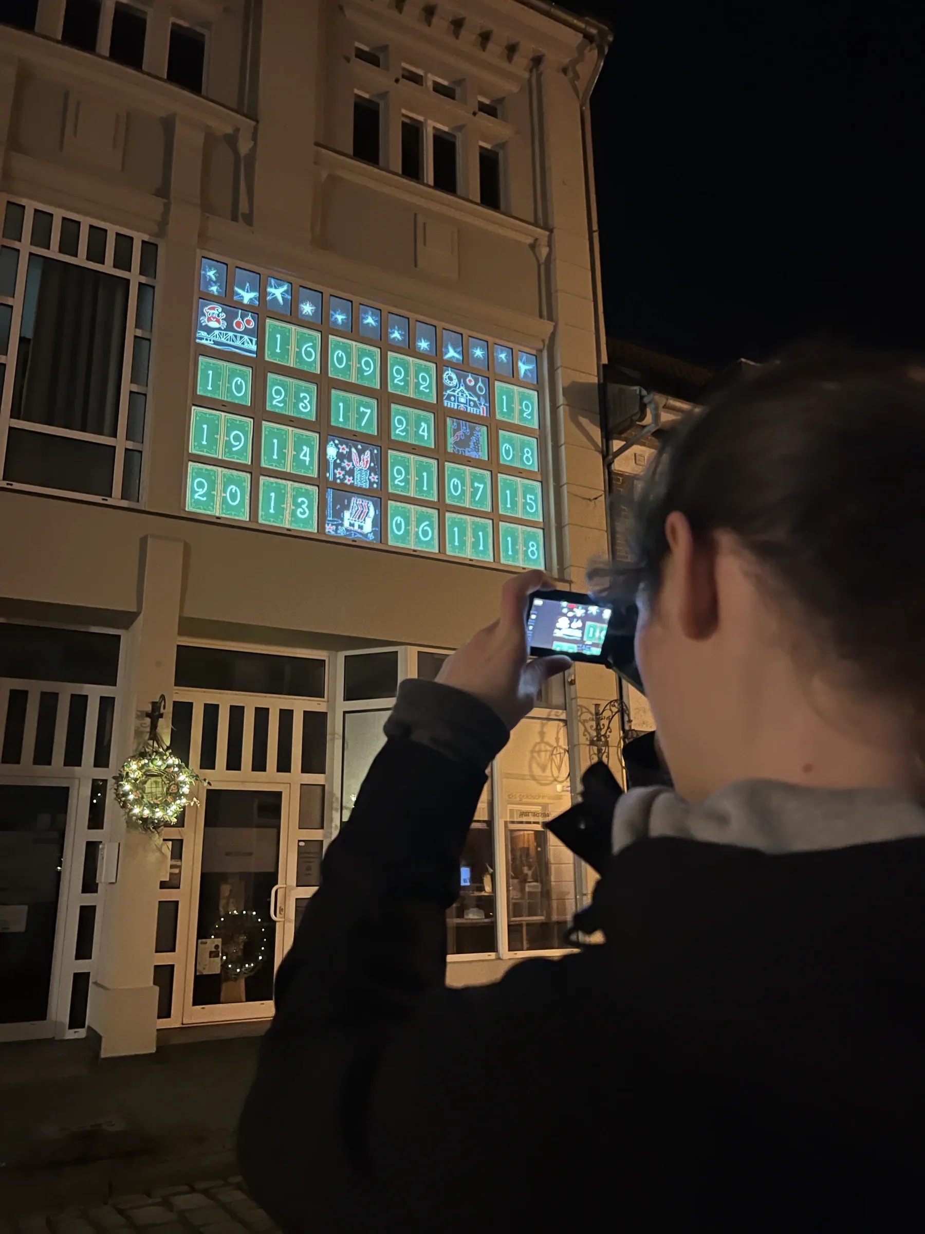 Woman photographing the façade in Barth, its windows showing a projected advent calendar with green numbered doors and the first opened motifs.